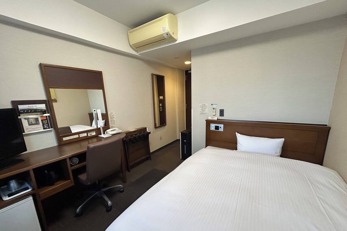 A hotel room with a bed, a desk with a computer, a chair, a mirror, and an air conditioning unit.