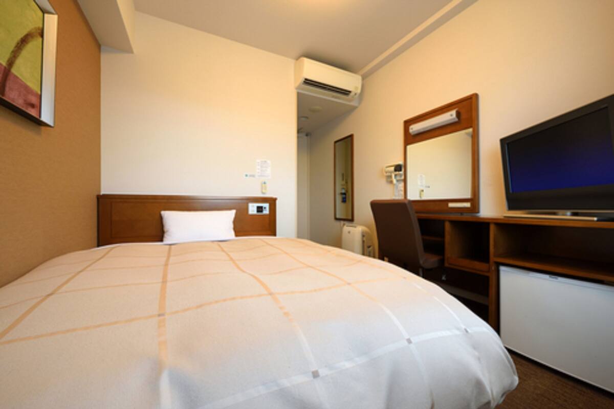 A hotel room with a large bed, a desk with a chair, a television, and an air conditioning unit.