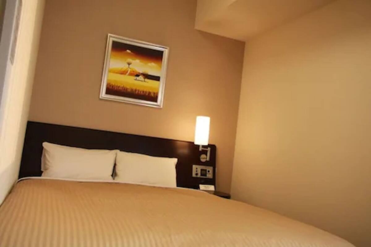 Quarto de hotel com cama, mesa de cabeceira, luminária na parede e uma pintura do pôr do sol.