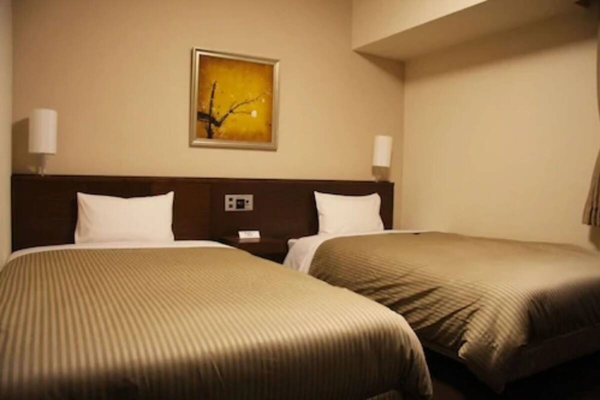 A hotel room with two beds, a nightstand, a wall-mounted painting, and two wall-mounted lamps.