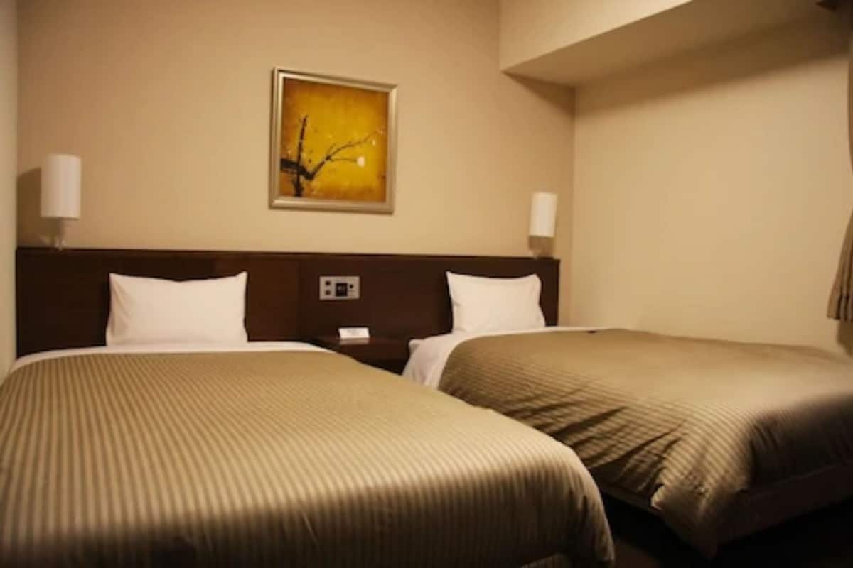 Quarto de hotel com duas camas, uma mesa de cabeceira, uma luminária na parede e um quadro.