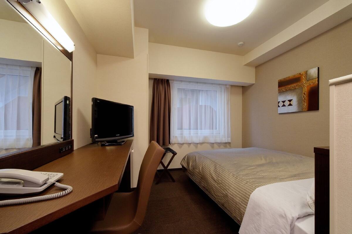 Une chambre d’hôtel avec un lit, un bureau avec un téléphone, une télévision et une fenêtre avec des rideaux.