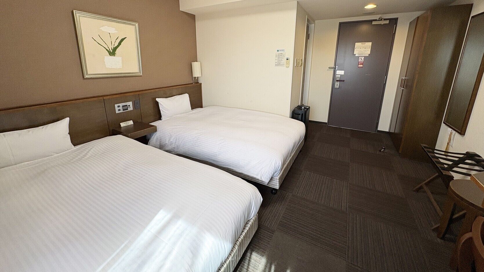 A hotel room with two beds, a bedside table, a lamp, a framed picture on the wall, a door, and a small table with a chair.