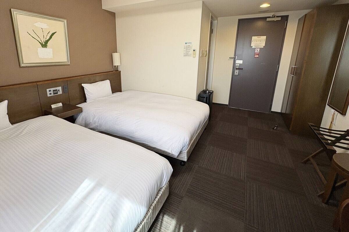 Une chambre d’hôtel avec deux lits, une table de chevet, une lampe, un tableau encadré au mur, une porte et une petite table avec une chaise.