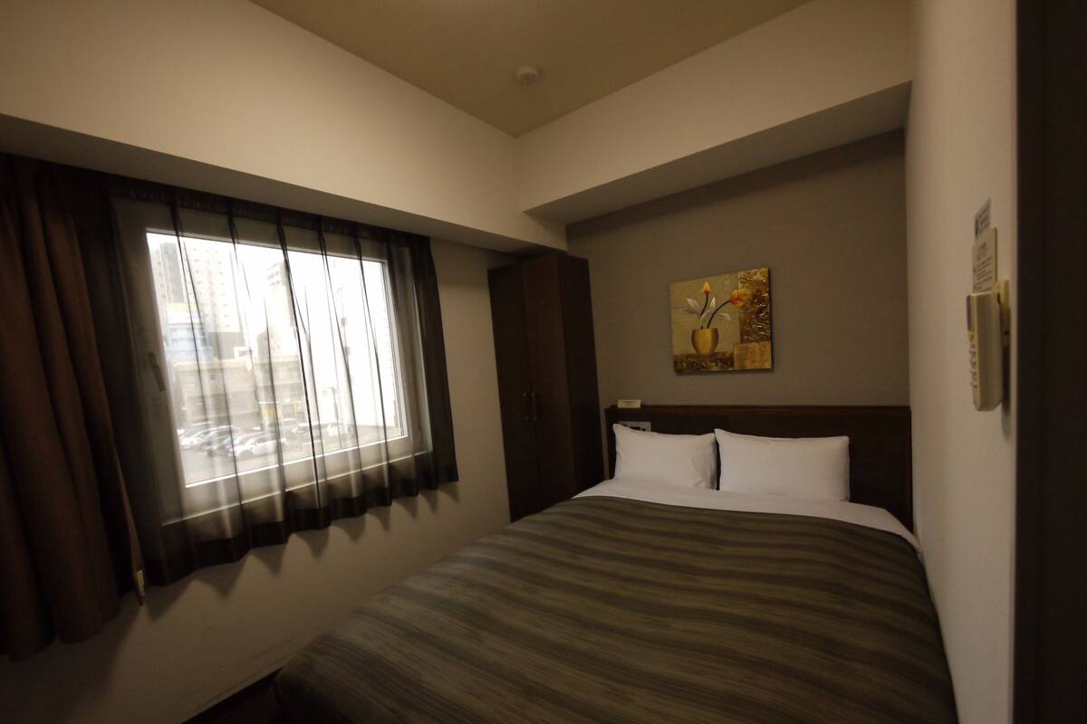 Une chambre d’hôtel avec un grand lit, une fenêtre avec des rideaux, un tableau au mur et un thermostat.
