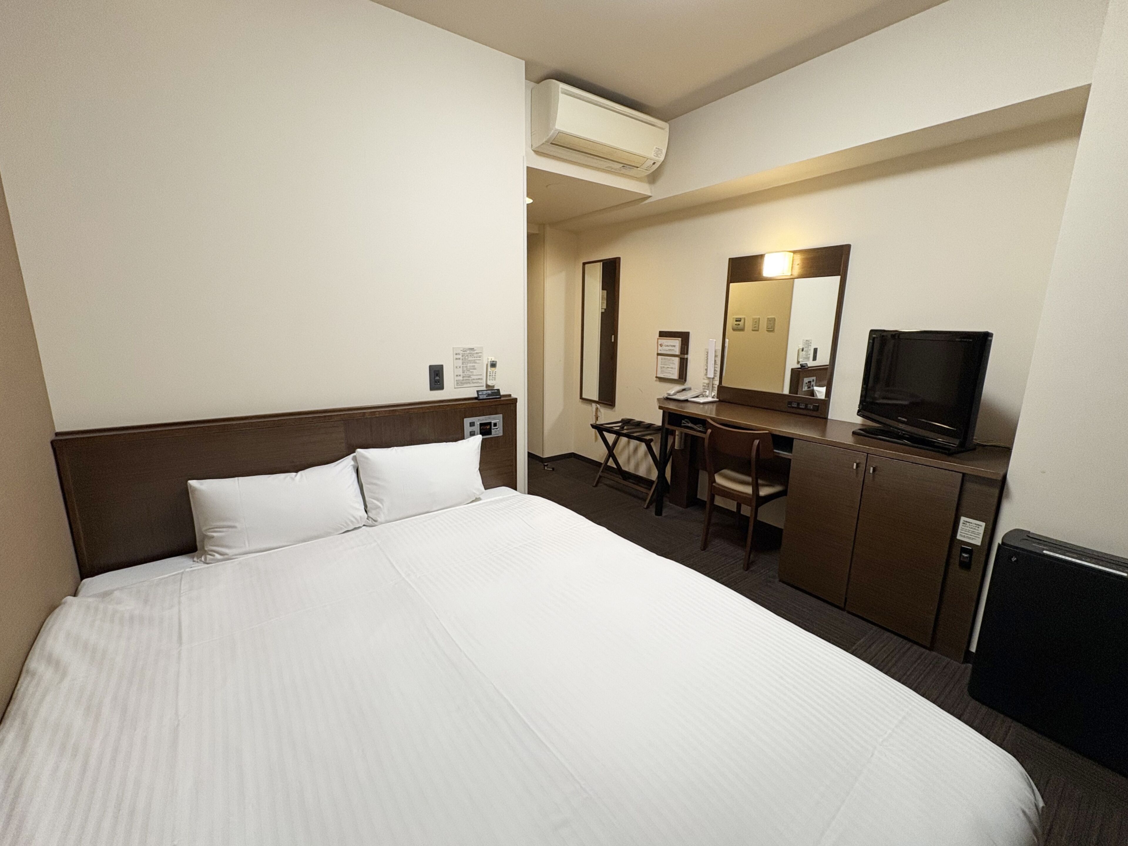 A hotel room with a bed, a desk, a chair, a television, and an air conditioning unit.