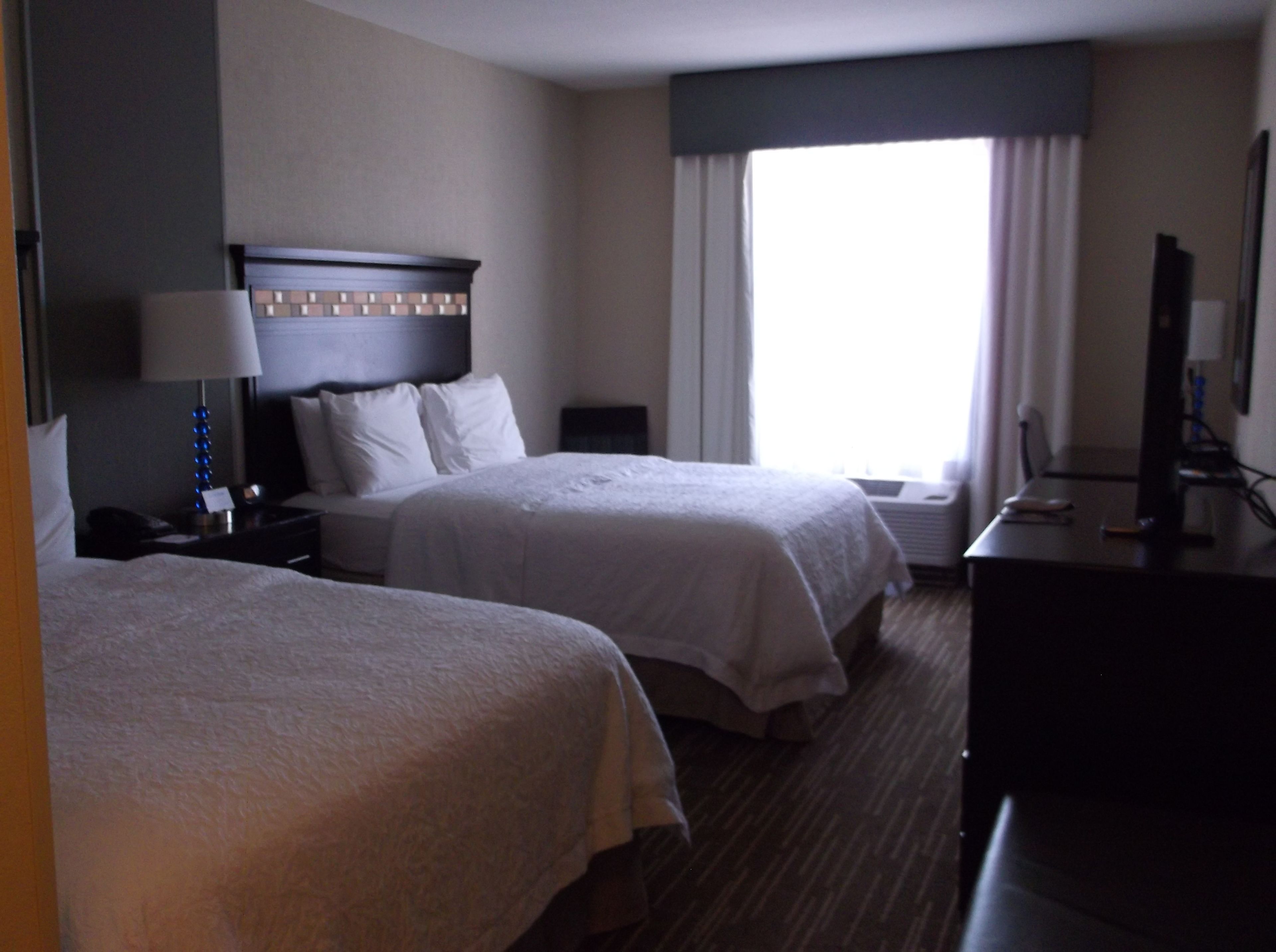 A hotel room with two beds, a desk, a lamp, and a large window with curtains.
