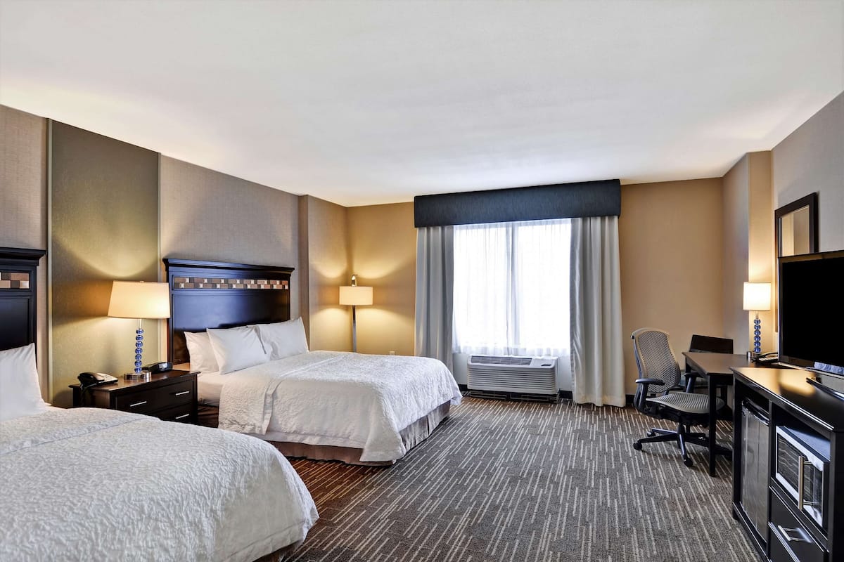 A hotel room with two beds, a desk, a chair, a TV, and a window with curtains.
