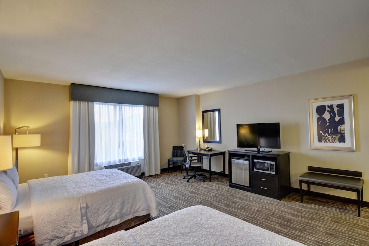 A hotel room with two beds, a desk with a television, a chair, a desk lamp, a bench, and a framed picture on the wall.