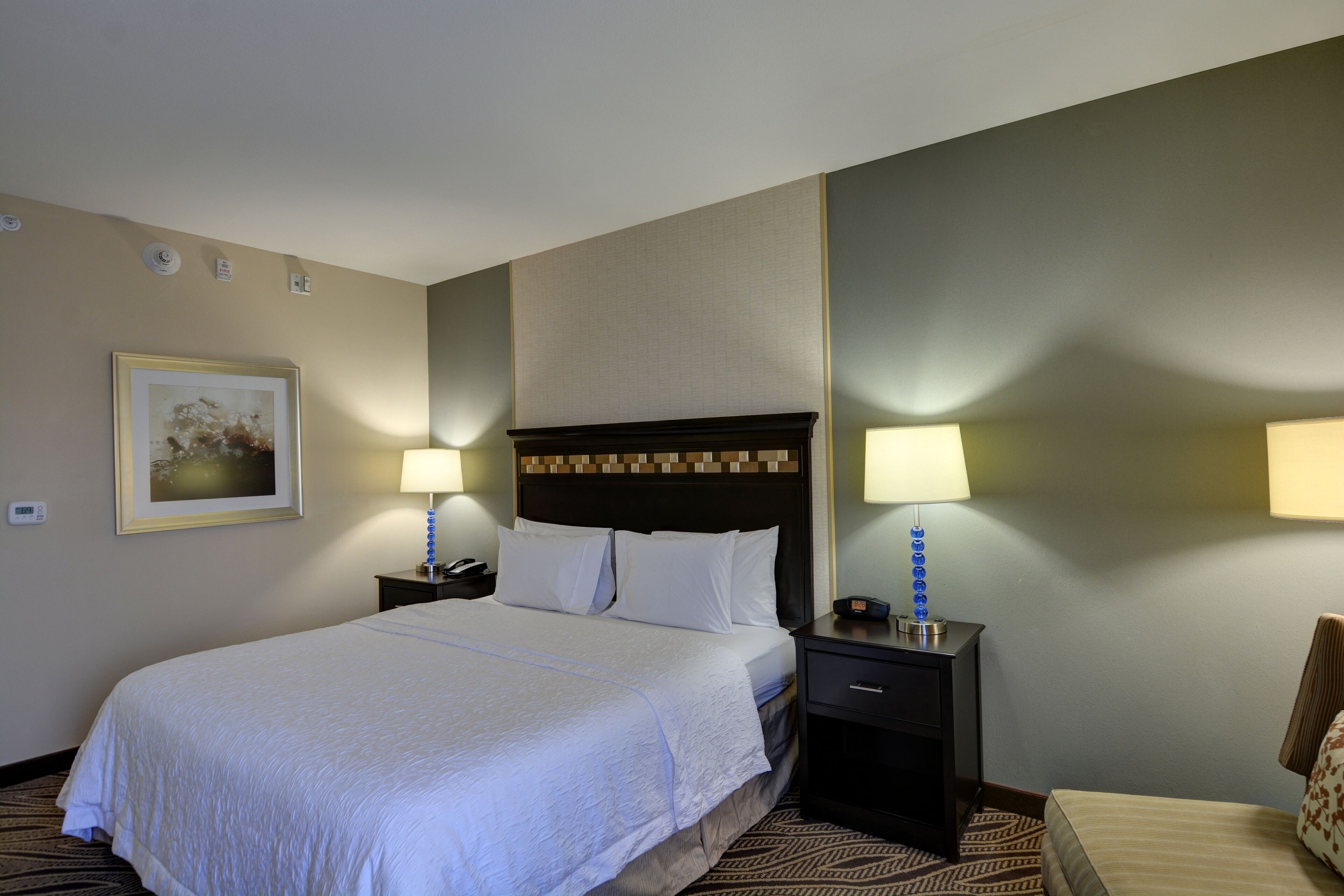 A hotel room with a large bed, two bedside tables with lamps, a framed picture on the wall, and a patterned carpet.