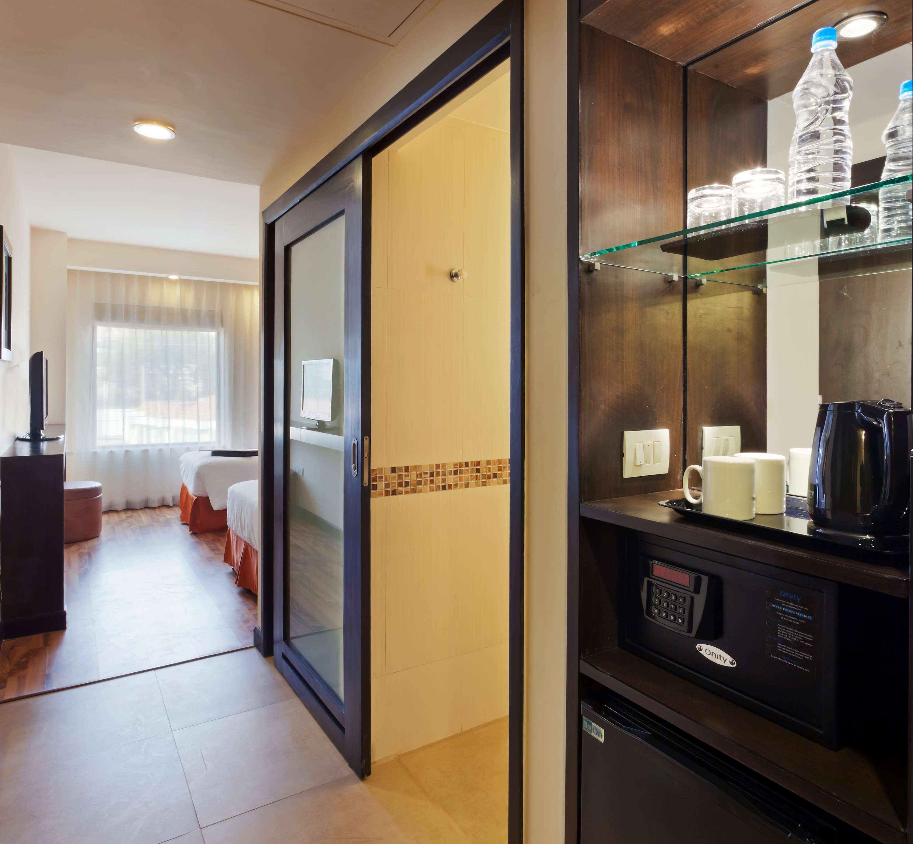A hotel room with a bed, a desk, a coffee machine, and a glass shelf with bottles.