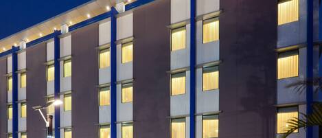 A modern hotel building with a prominent Hampton sign, multiple windows, and a well-lit facade.