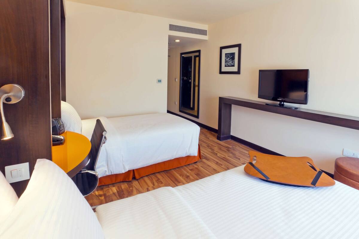 Quarto de hotel com duas camas, uma escrivaninha, uma televisão e uma porta.