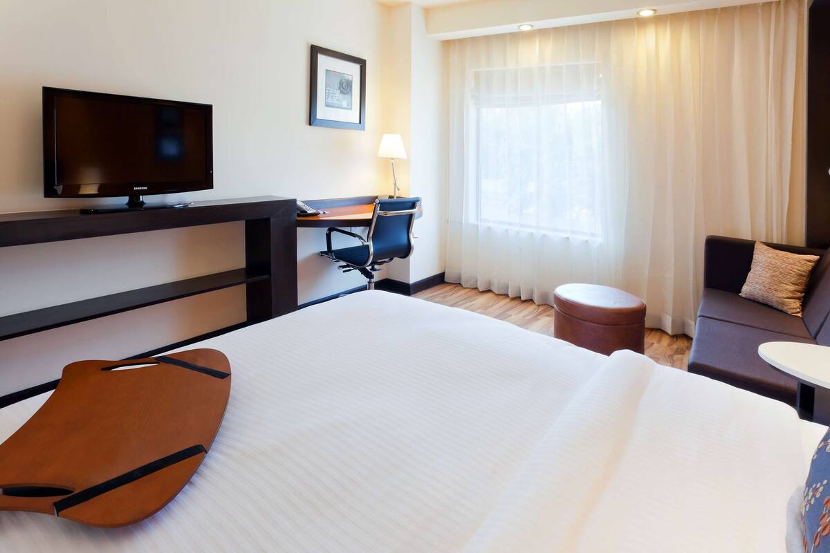 Quarto de hotel com cama, escrivaninha com cadeira, televisão, janela com cortinas e sofá.