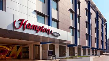 A modern hotel exterior with a prominent "Hampton by Hilton" sign, large windows, and a clean, contemporary design.