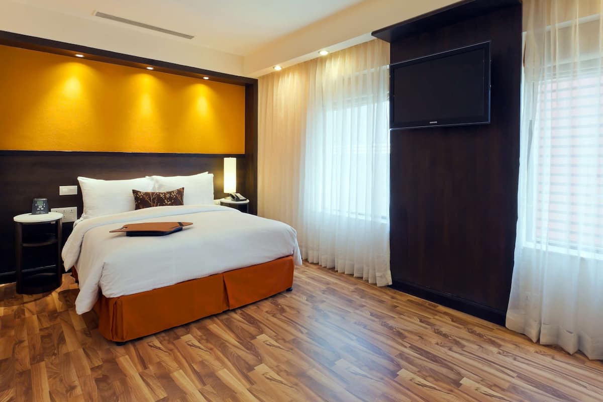 Quarto de hotel com uma cama grande, televisão de tela plana, mesa de cabeceira com abajur e piso de madeira.