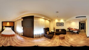 Vista panorámica de una habitación de hotel con piso de madera curva, una cama, un televisor, un sofá, un escritorio y una silla.