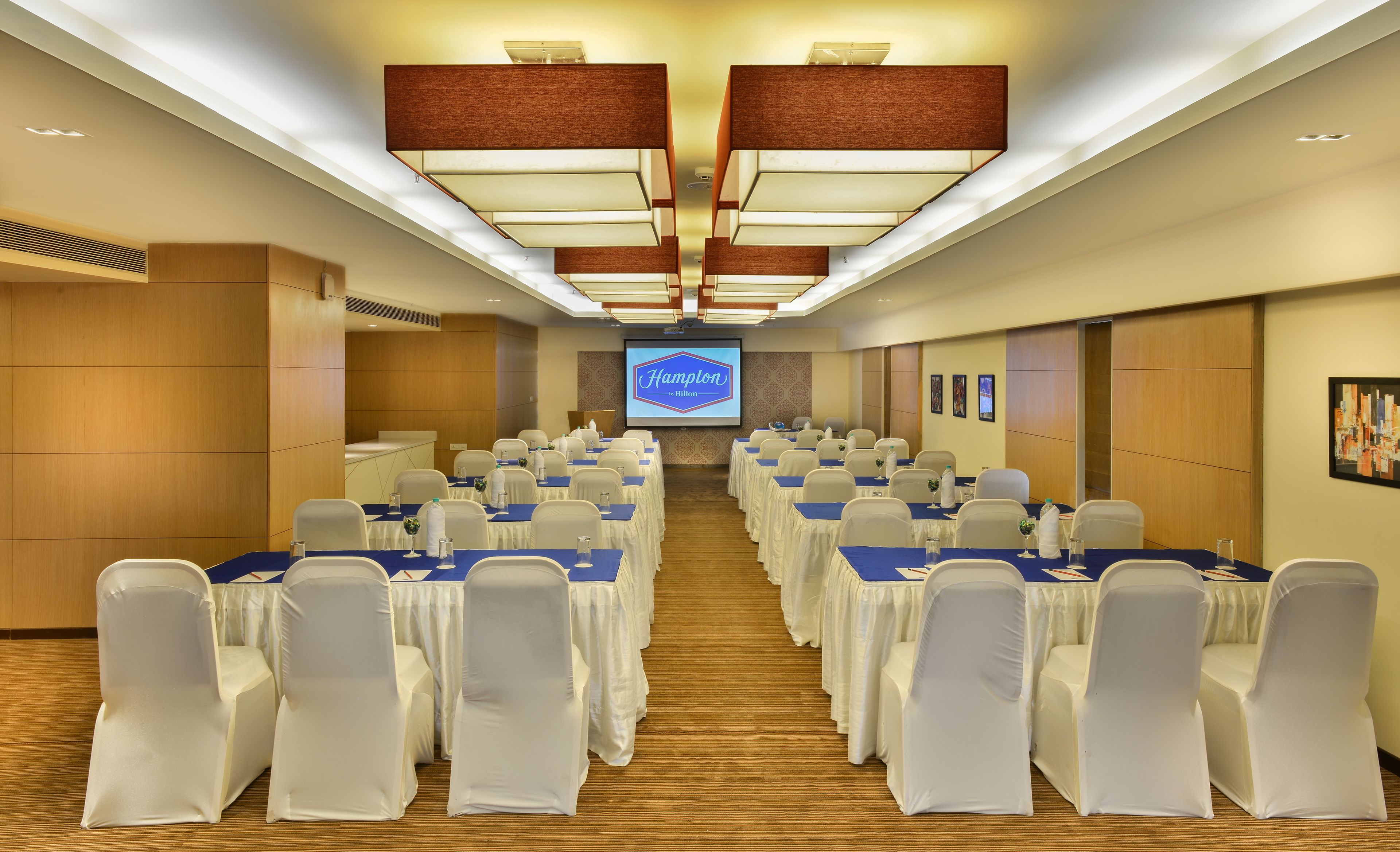 A conference room with rows of tables and chairs, a large screen displaying 'Hampton by Hilton', and wooden panel walls.