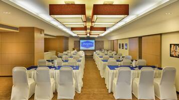 A conference room with rows of tables and chairs, a large screen displaying 'Hampton by Hilton', and wooden panel walls.