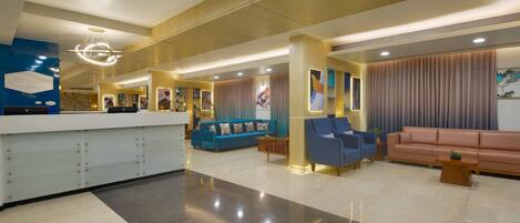 A hotel lobby with a reception desk, blue and pink seating areas, and a modern chandelier.