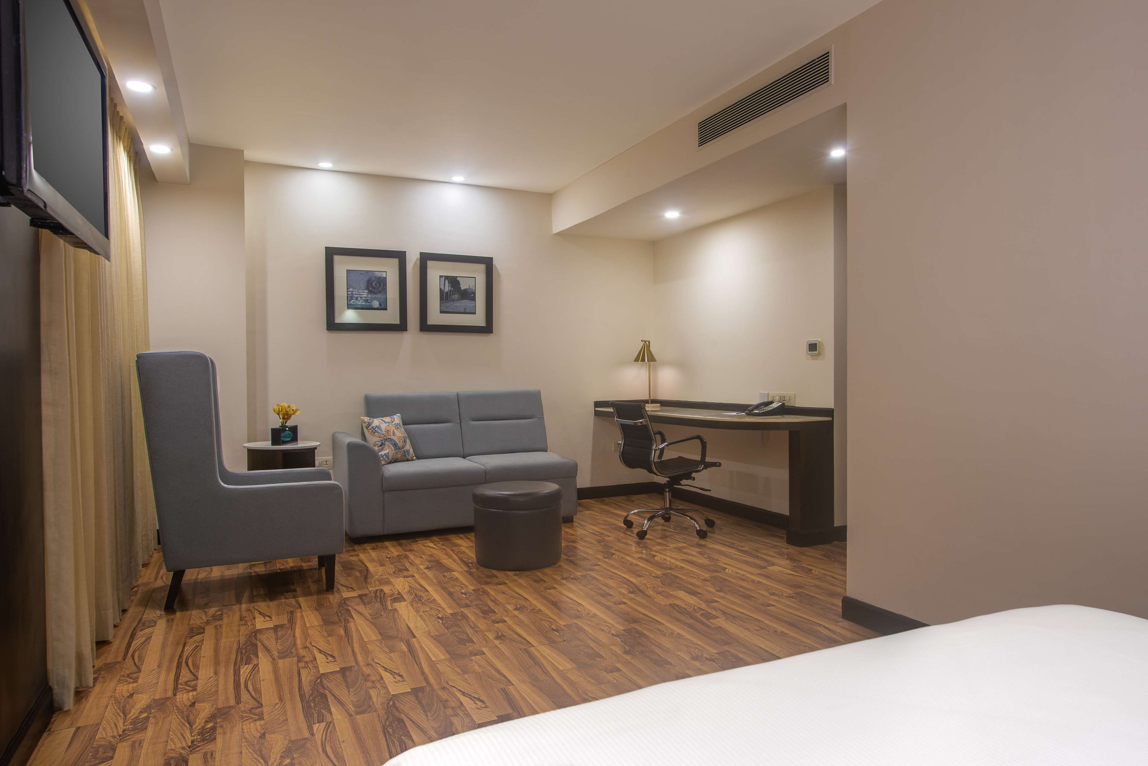 Una habitación de hotel con una cama, un sillón gris, una mesita con un florero, un sofá gris, un escritorio con una silla y dos cuadros en la pared.