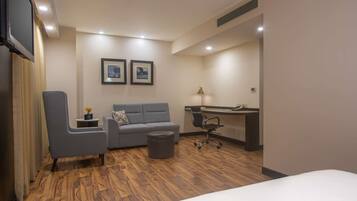 Una habitación de hotel con una cama, un sillón gris, una mesita con un florero, un sofá gris, un escritorio con una silla y dos cuadros en la pared.