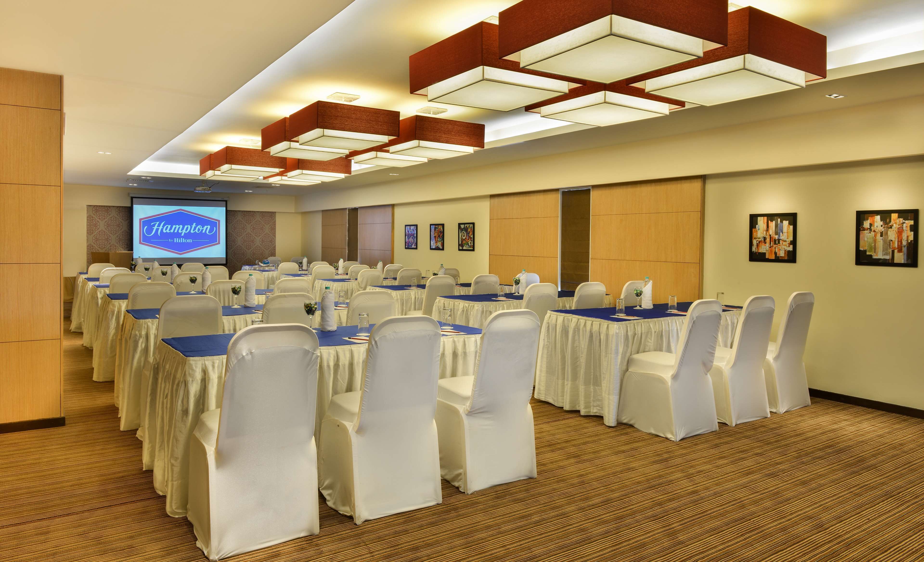 A conference room with rows of tables and chairs, a projector screen, and artwork on the walls.