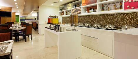 A modern restaurant with white tables and chairs, a buffet counter with food items, and shelves with bottles and glasses.