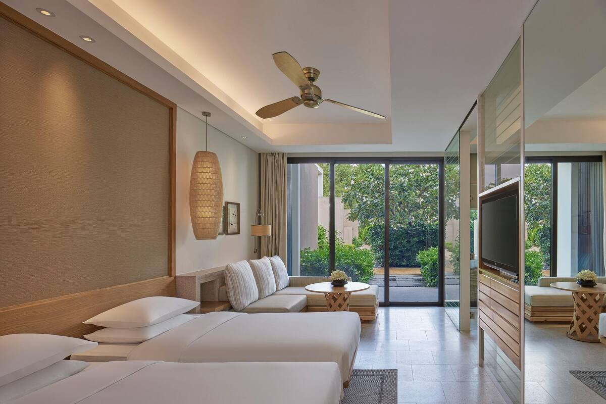A modern hotel room with a large bed, a sofa, a TV, and a balcony with a view of greenery.