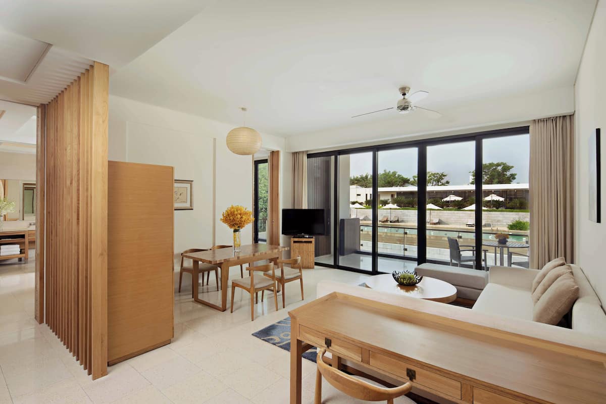 A modern hotel room with a dining area, a sofa, a TV, and large windows overlooking a pool area.