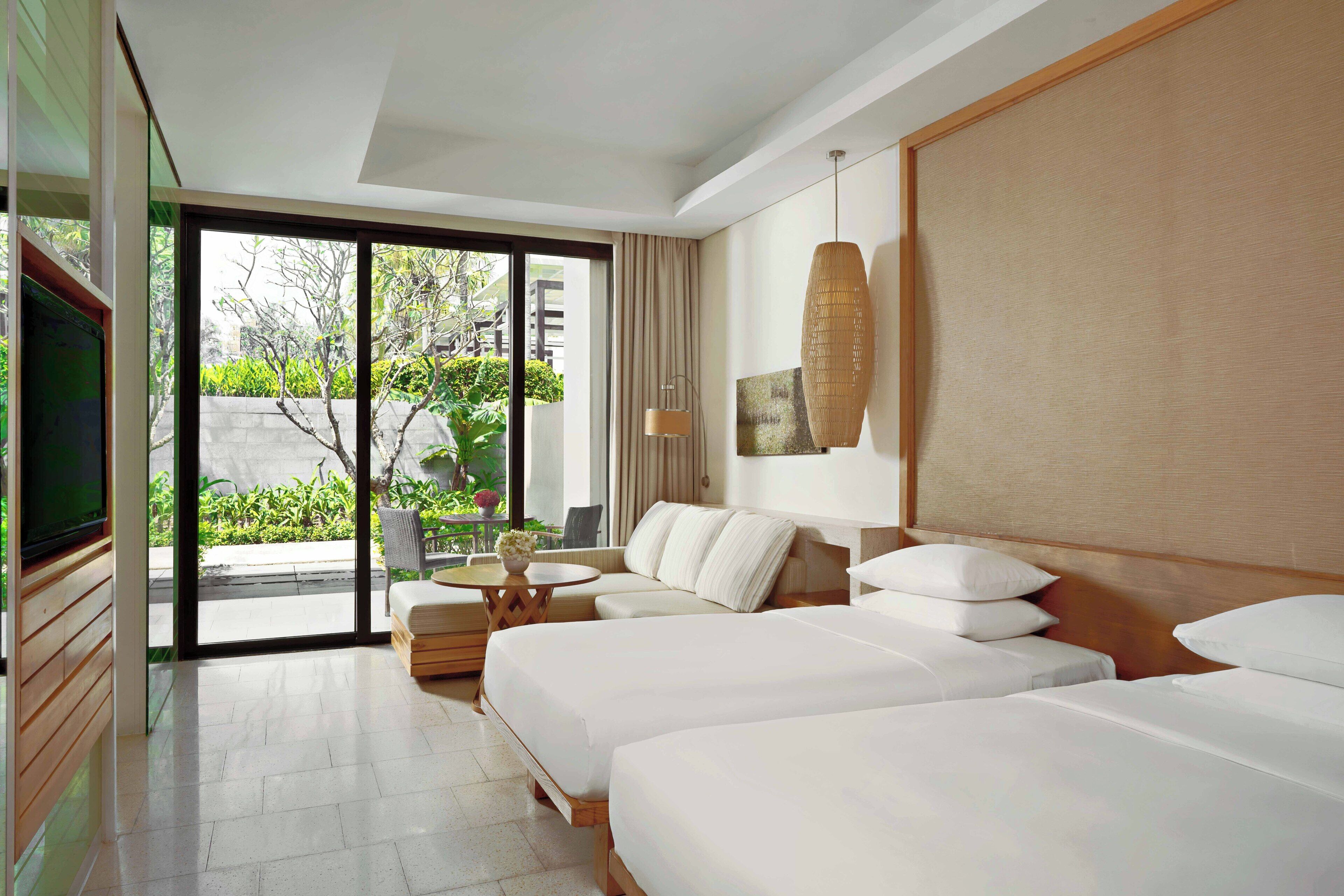A hotel room with two beds, a sofa, a TV, and a balcony with a view of greenery.