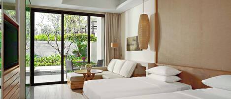 A hotel room with two beds, a sofa, a TV, and a balcony with a view of greenery.