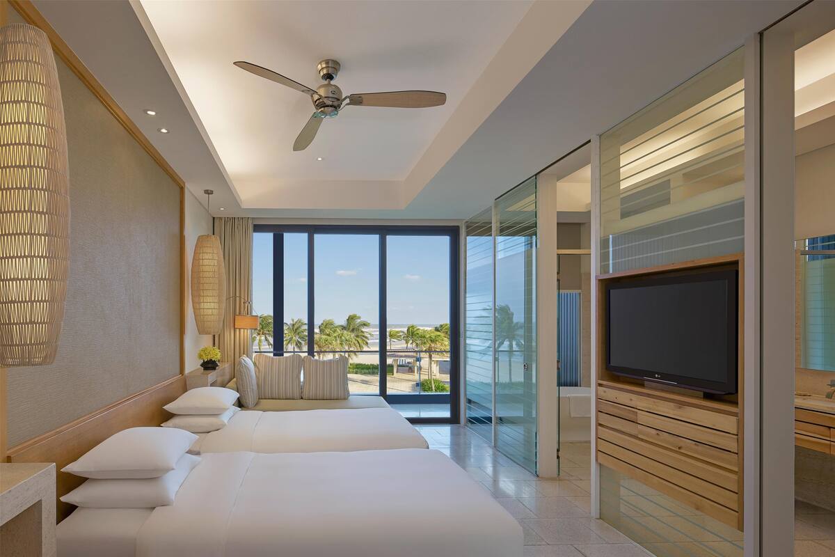 A hotel room with a large bed, a ceiling fan, a TV, and a view of the beach.
