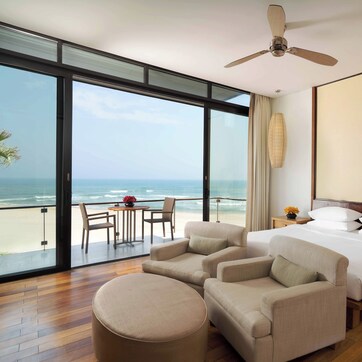 A hotel room with a large window overlooking the beach, a bed, two armchairs, a small table, and a ceiling fan.