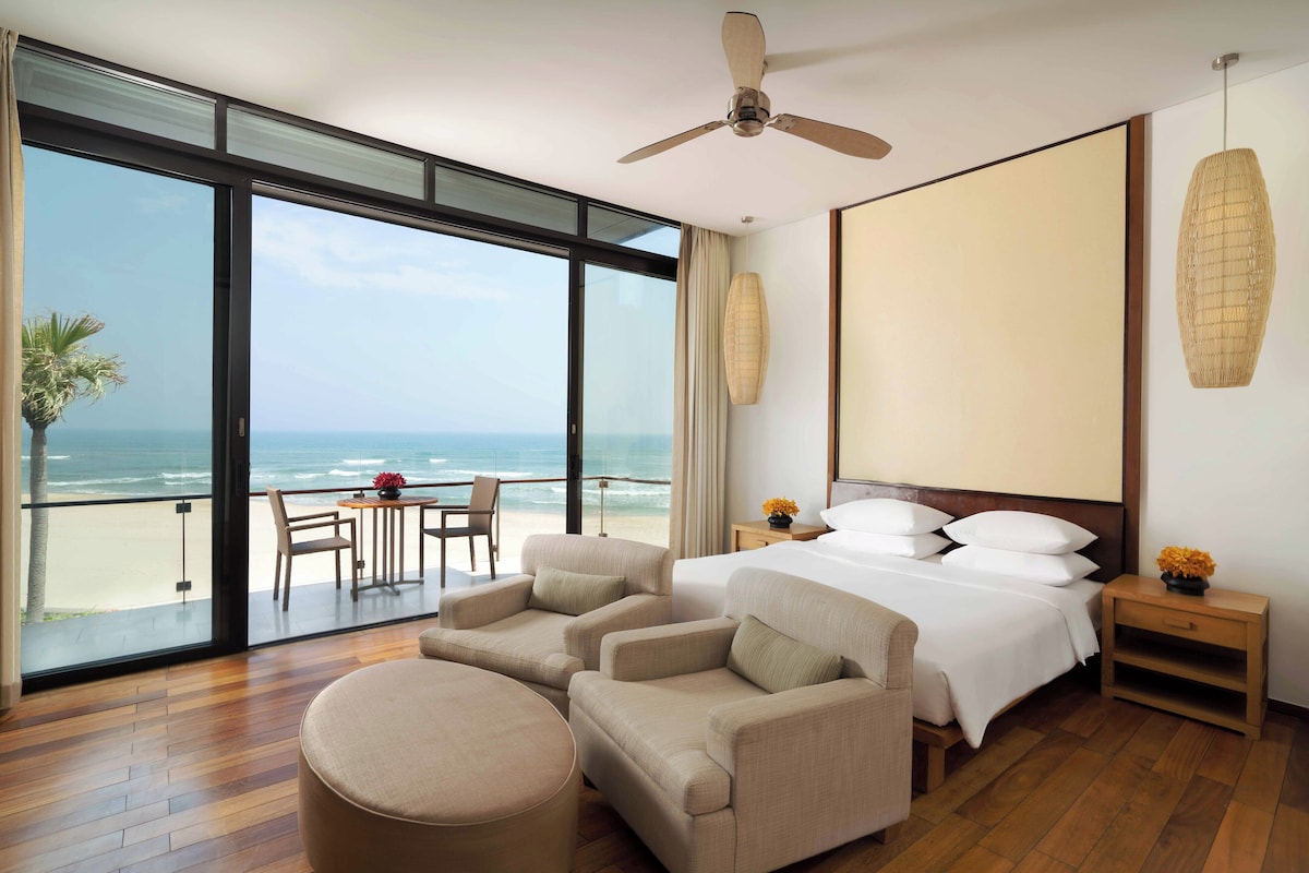 A hotel room with a large window overlooking the beach, a bed, two armchairs, a small table, and a ceiling fan.