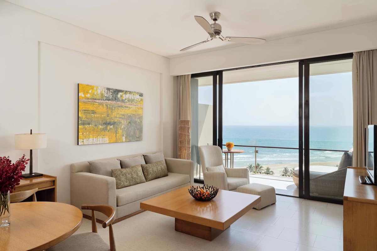 A modern living room with a sofa, coffee table, and chairs, a painting on the wall, and a view of the ocean through large windows.