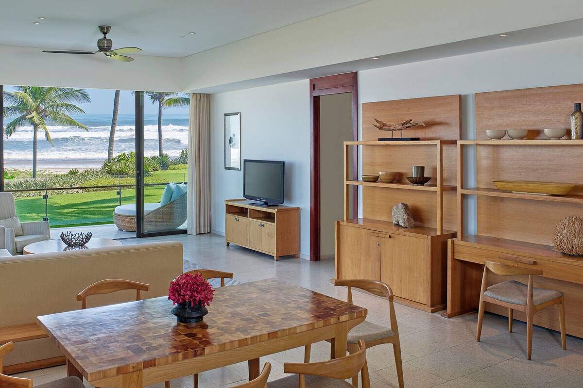 A modern living room with a wooden dining table, chairs, a TV, and a view of the ocean.