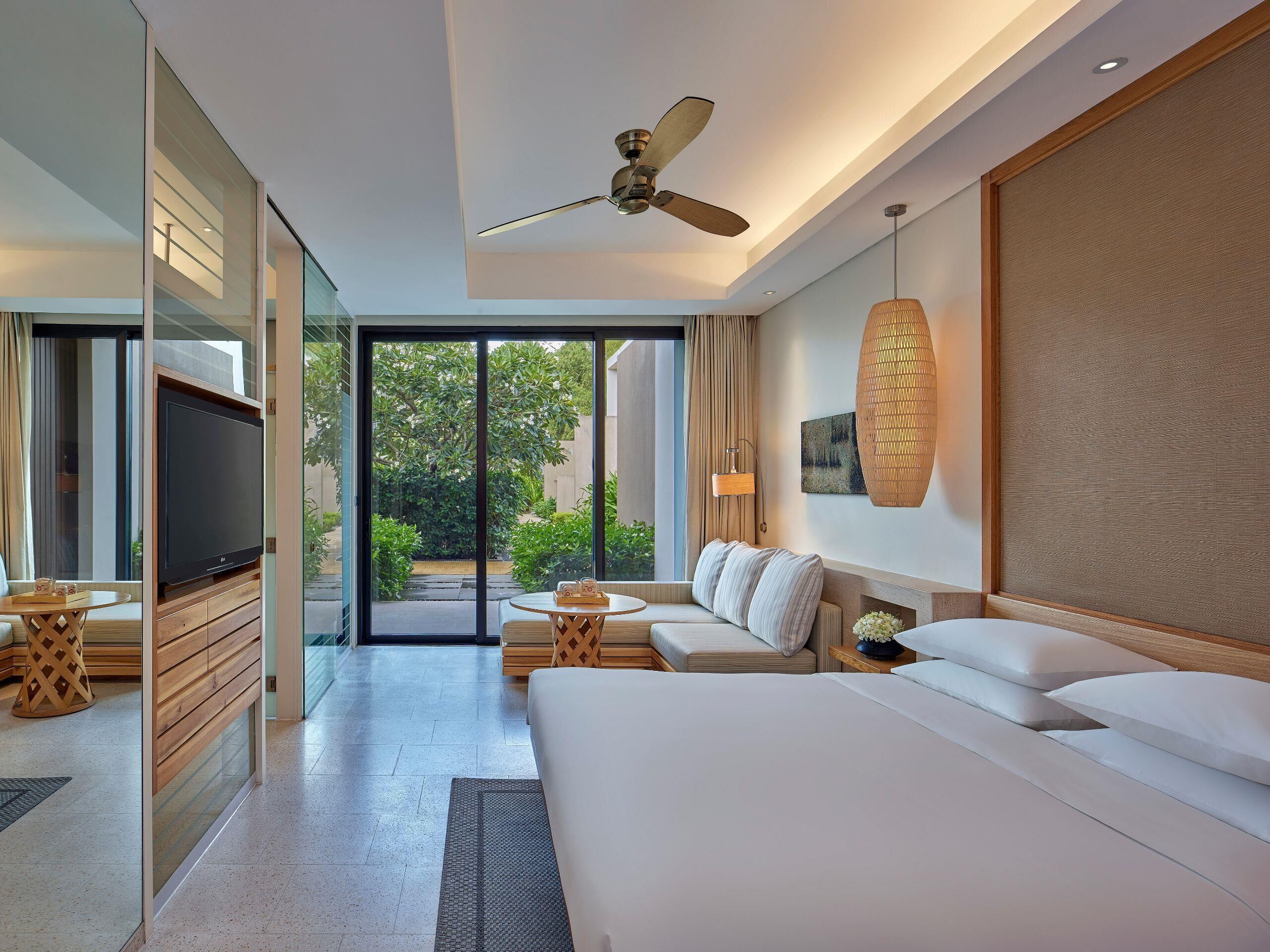 A modern hotel room with a large bed, a sofa, a TV, and a balcony with a view of greenery.