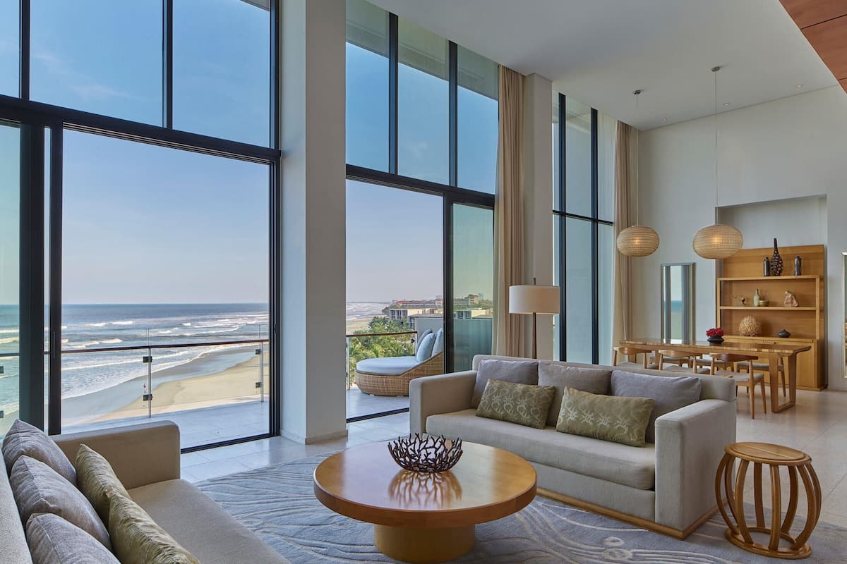 A spacious living room with a large window overlooking a beach, a sofa, a coffee table, and a dining area with a wooden table and chairs.