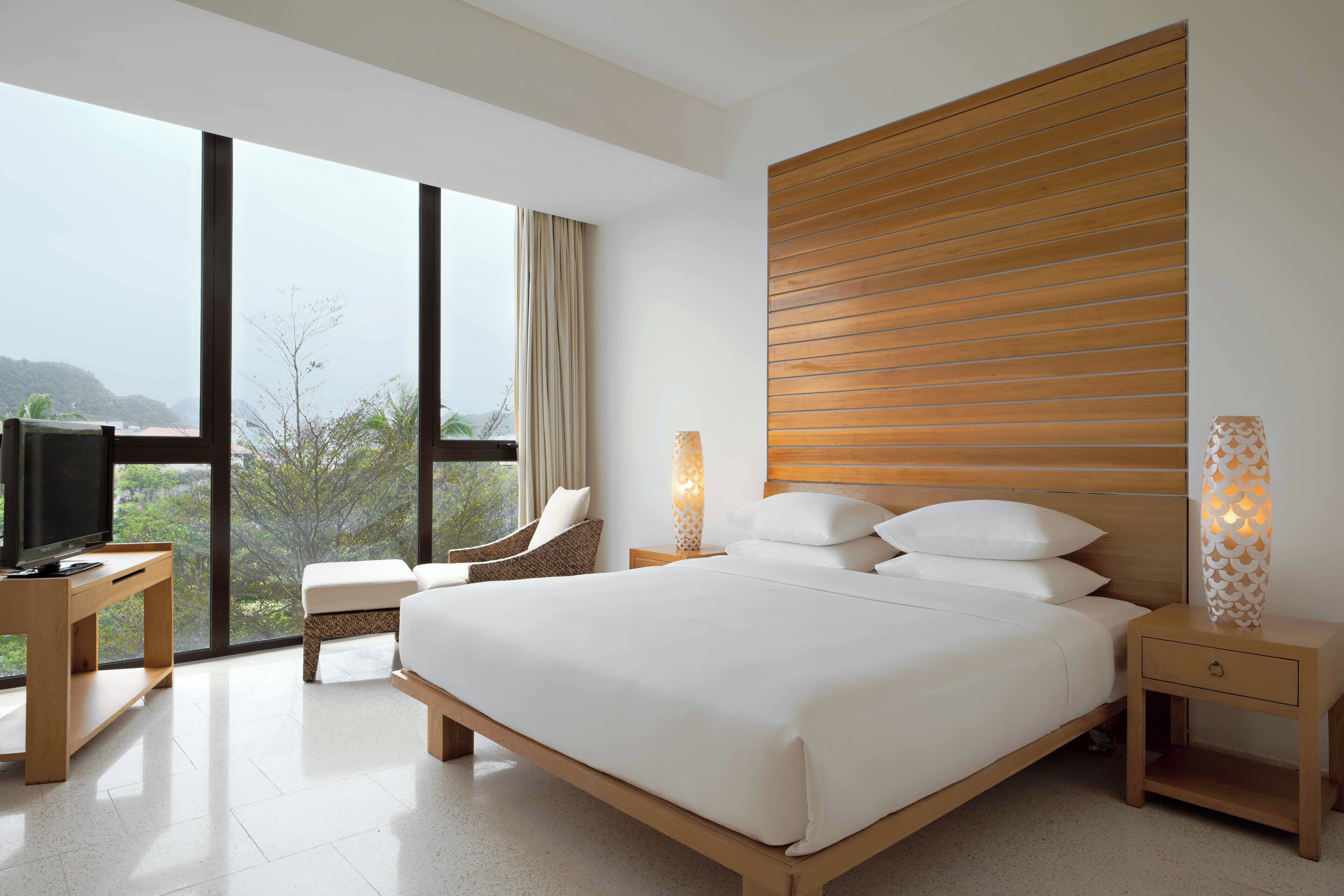 Ein modernes Hotelzimmer mit einem großen Bett, einem Fernseher auf einem Holztisch, Blick ins Freie und einem Holz-Kopfschild.