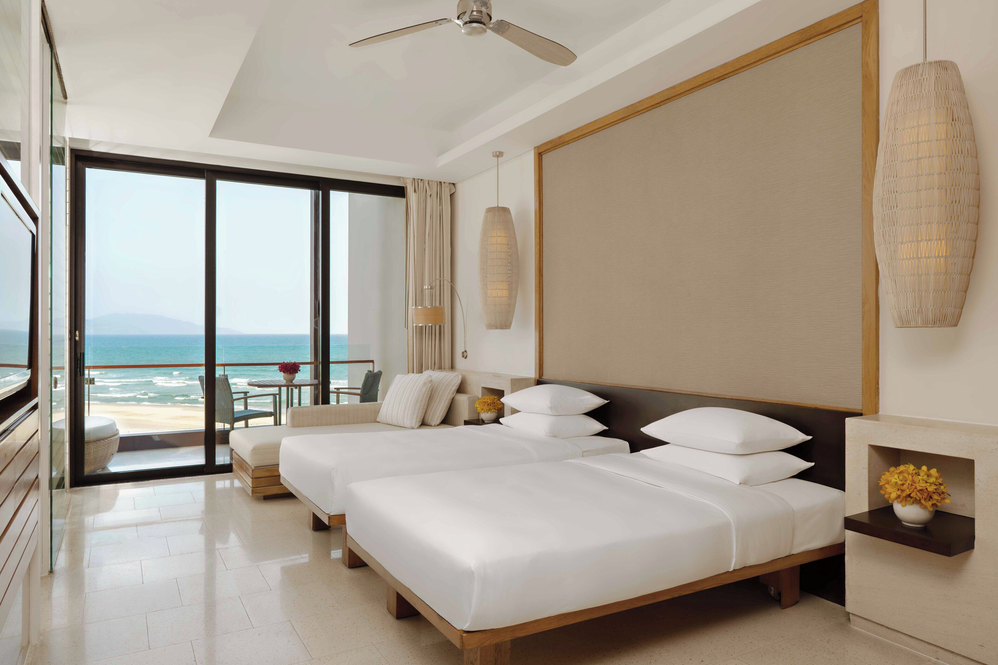 A modern hotel room with two beds, a balcony with a view of the ocean, a ceiling fan, and a large vase of flowers on the nightstand.