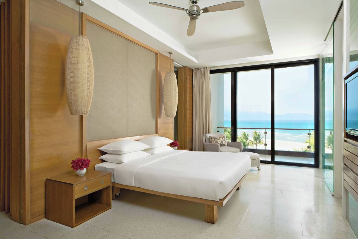 A modern hotel room with a large bed, wooden headboard, a bedside table with a vase of flowers, a ceiling fan, and a view of the ocean through large windows.