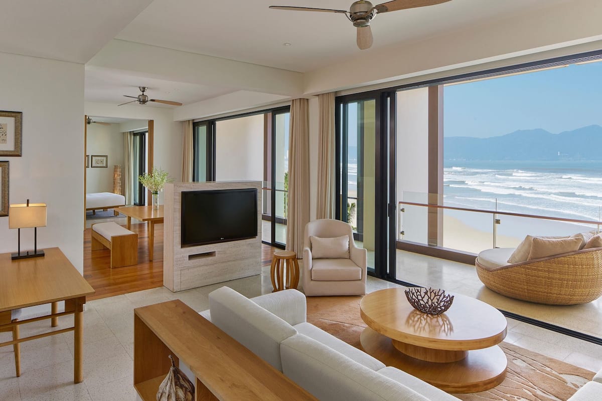 A modern living room with a flat-screen TV, comfortable seating, a dining area, and a balcony with a view of the ocean.