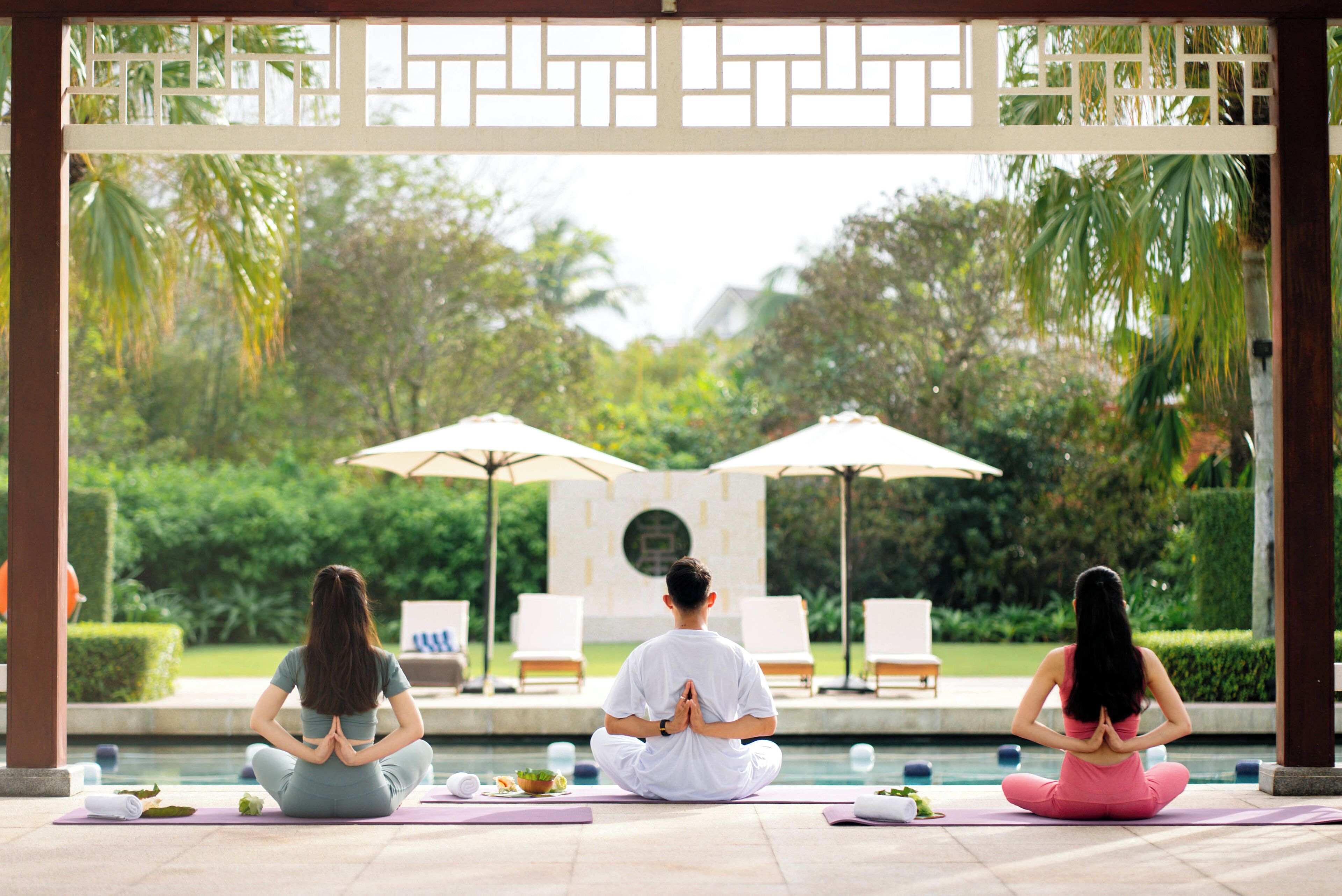 Three individuals meditating by a poolside with umbrellas and lounge chairs in the background.