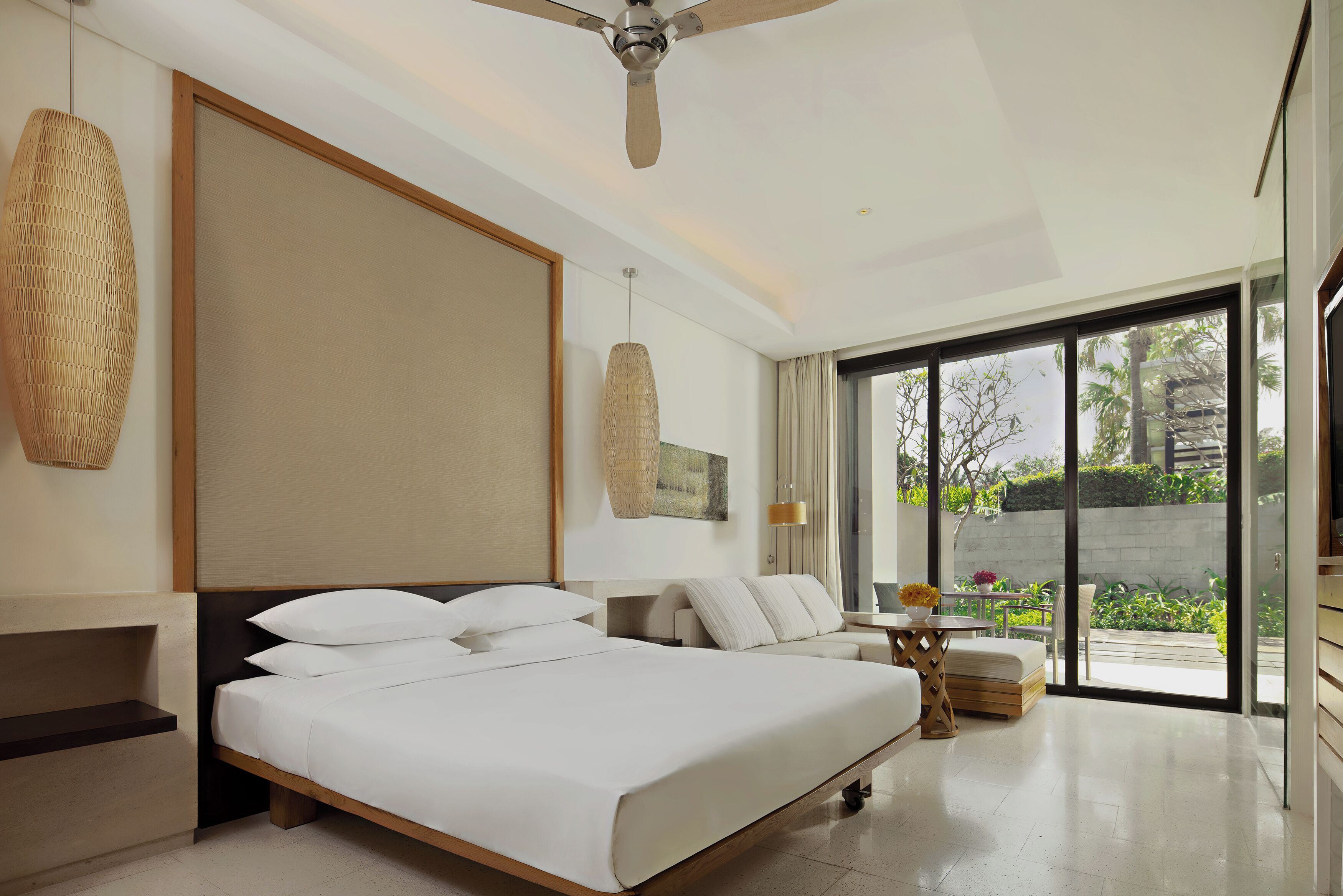A modern hotel room with a large bed, a ceiling fan, a view of the outdoors, and a small seating area with a table and chairs.