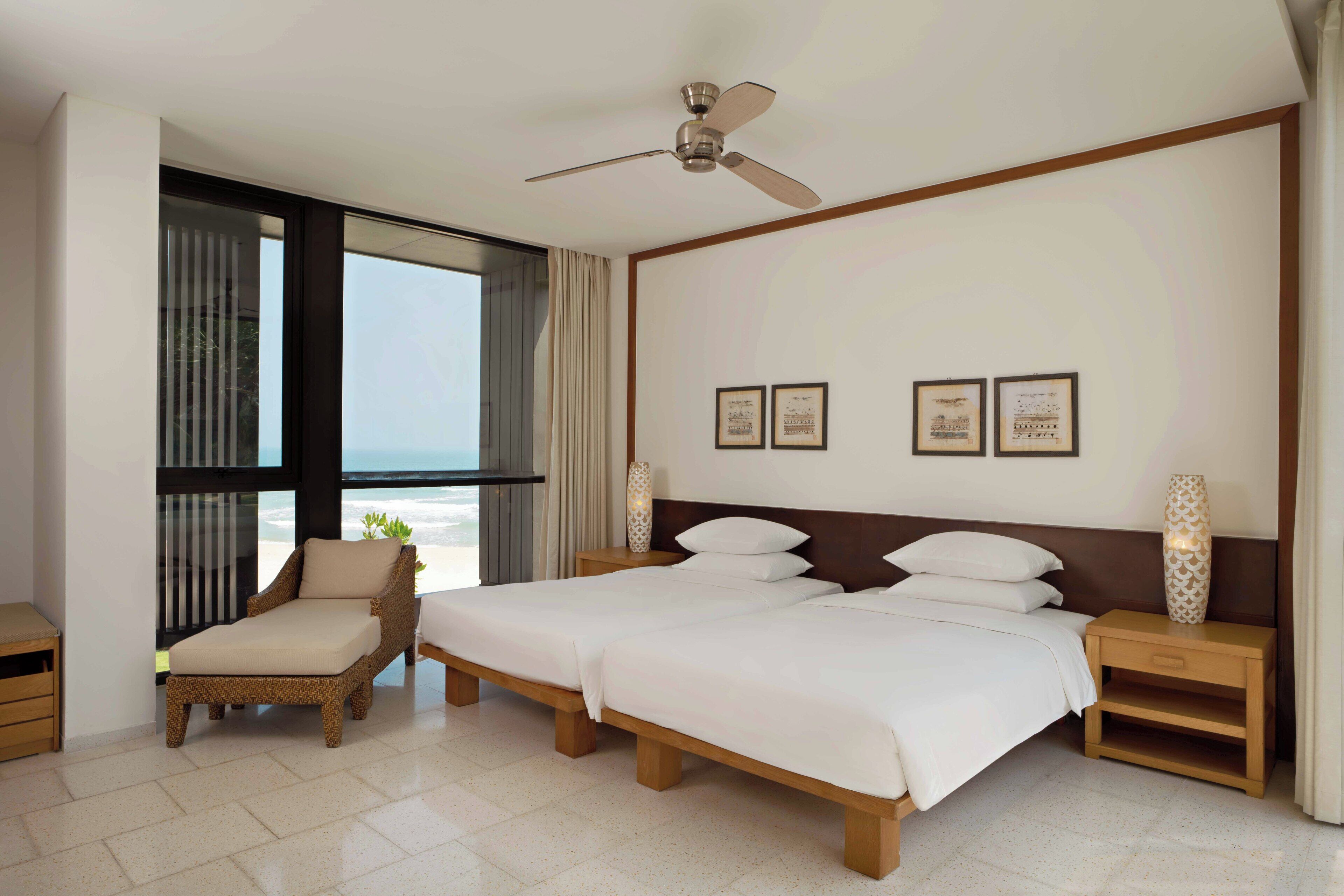 Habitación de hotel con una cama grande, una silla, una mesita de noche y un ventilador de techo. En la pared hay diplomas enmarcados y una ventana con cortinas.