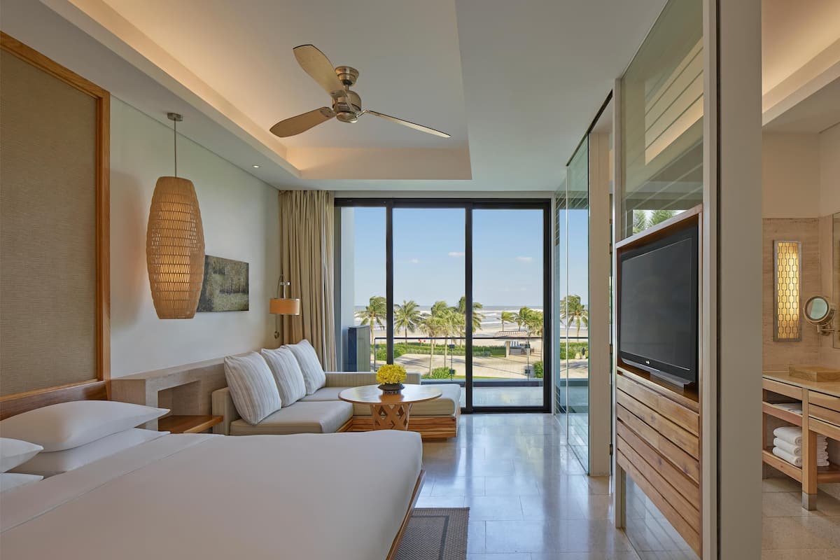 A hotel room with a large bed, a TV, a ceiling fan, and a view of the beach.