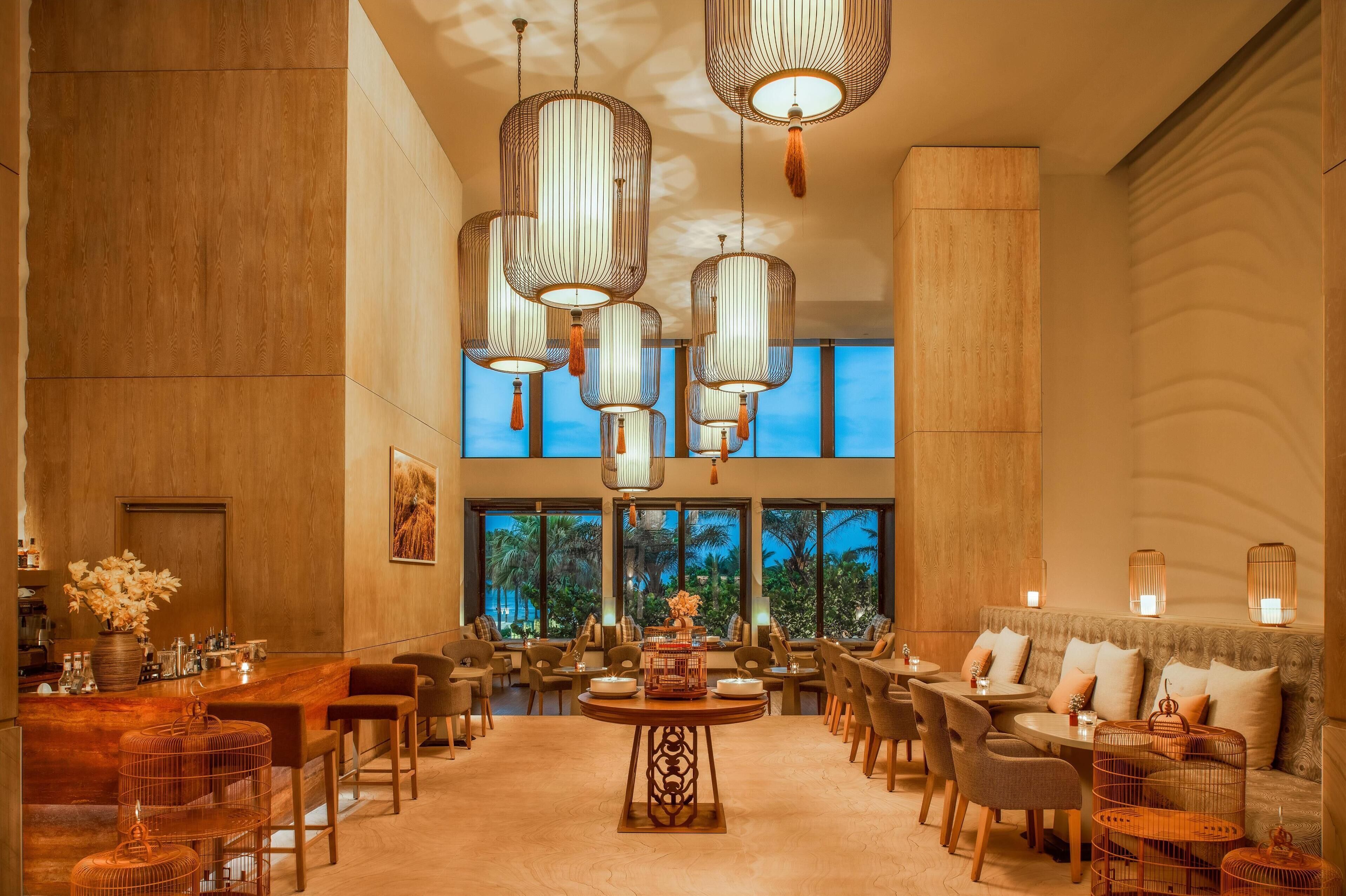 Un hall d’hôtel spacieux, doté d’une grande table ronde au centre, de plusieurs espaces aménagés pour s’asseoir et de grandes fenêtres offrant une vue sur les palmiers.