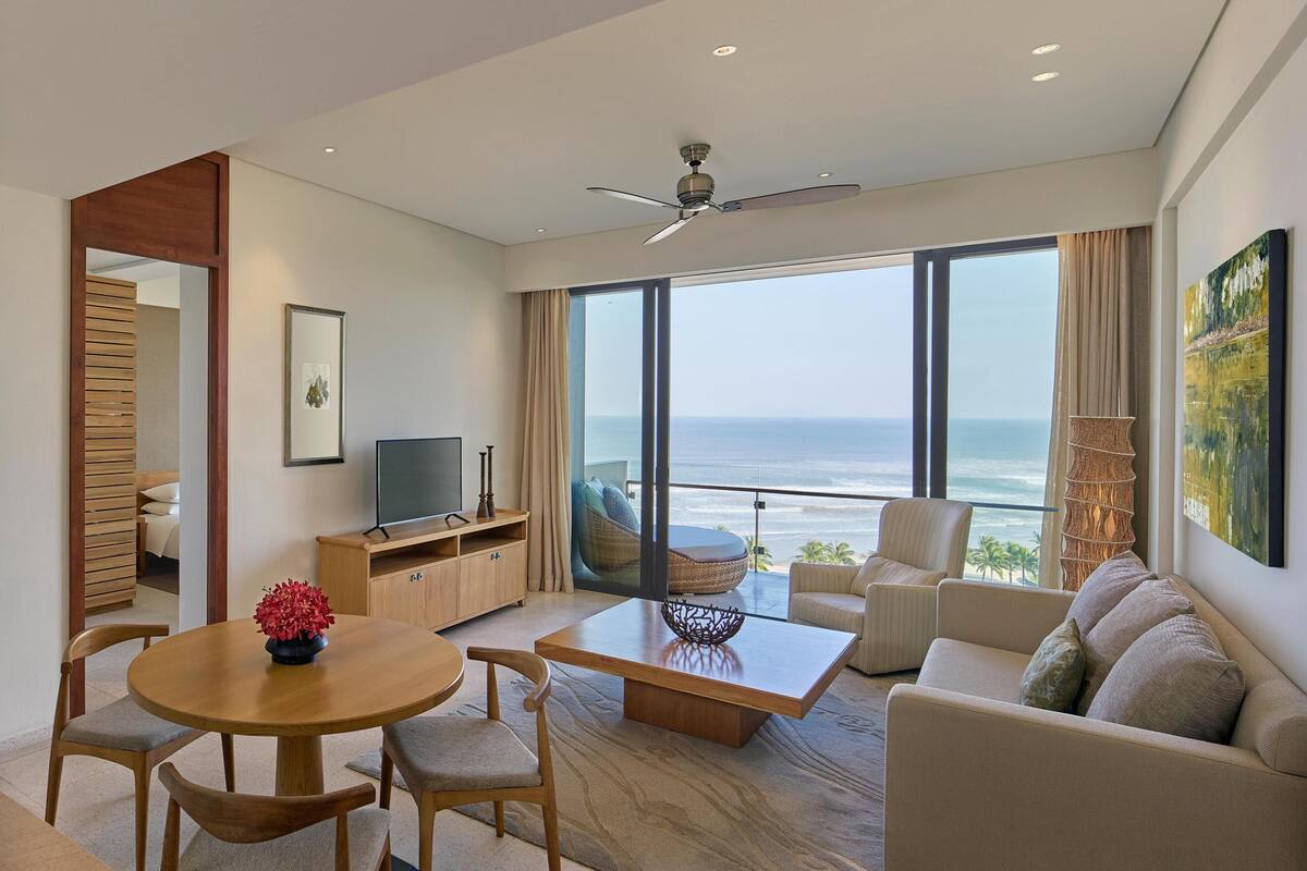 A modern living room with a large window overlooking the ocean, a ceiling fan, and a dining area with a table and chairs.