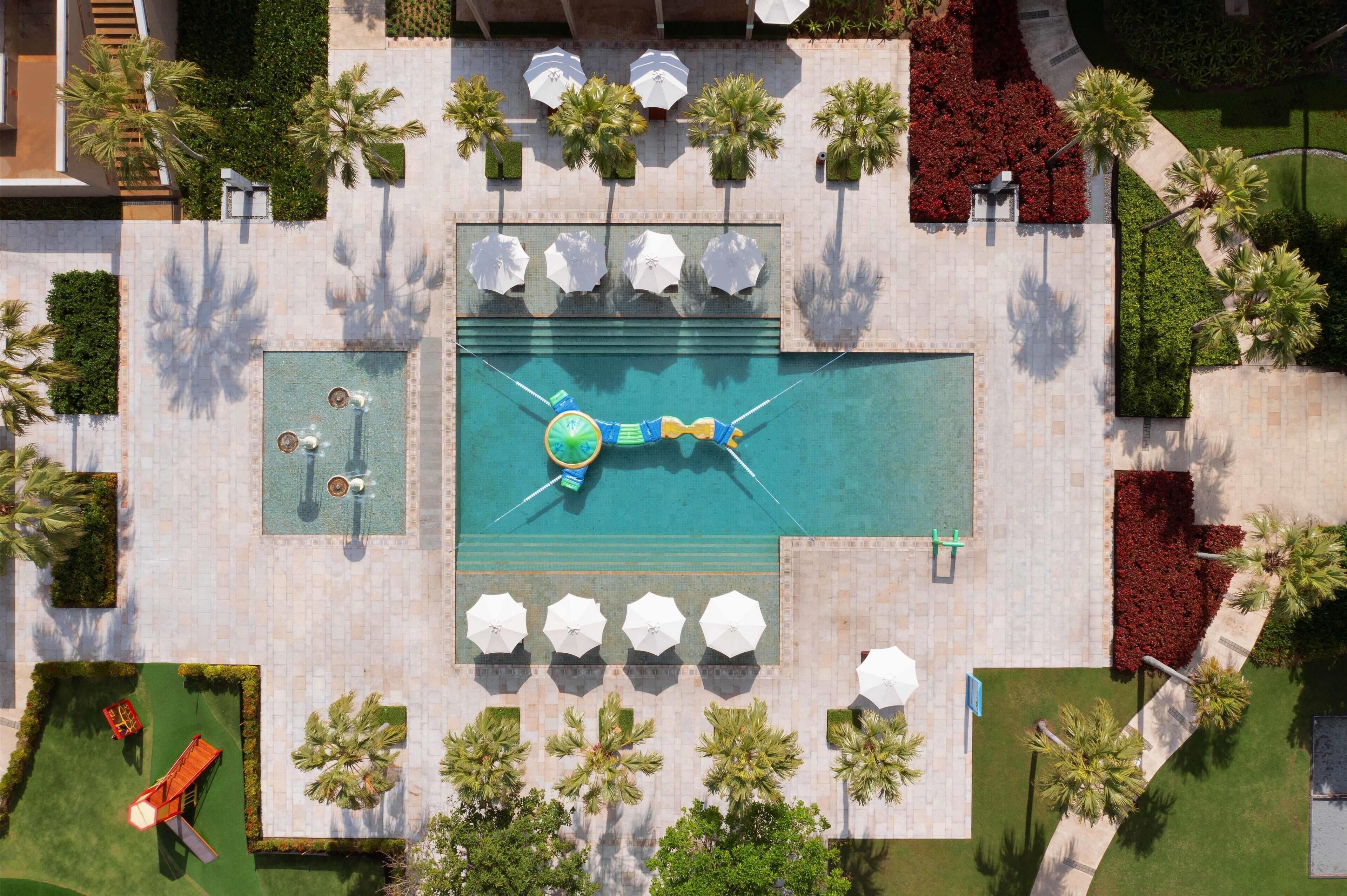 An aerial view of a resort with a central swimming pool, multiple lounge areas with umbrellas, and surrounding landscaped gardens.